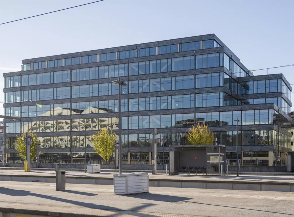 En modern glasbyggnad med reflekterande fasad, omgiven av en öppen gångväg och växtlighet.