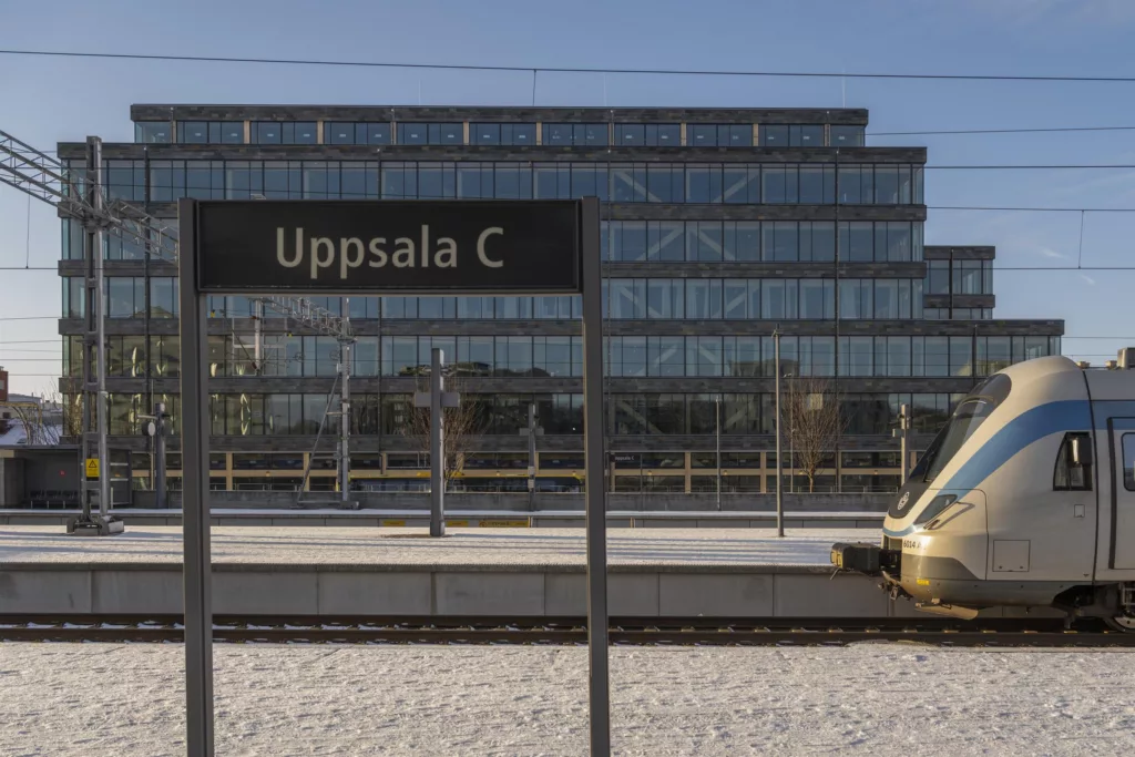 En skylt med texten "Uppsala C" framför ett glasbeklätt byggnadskomplex, med järnvägsspår och ett guldfärgat tåg i förgrunden.