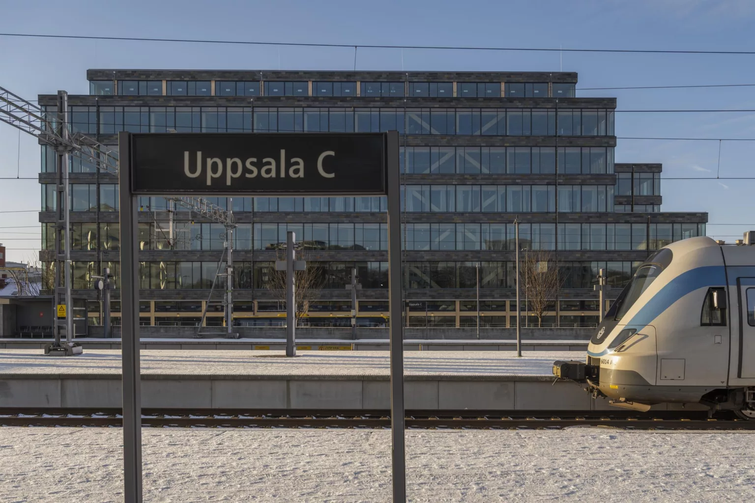 En skylt med texten "Uppsala C" framför ett glasbeklätt byggnadskomplex, med järnvägsspår och ett guldfärgat tåg i förgrunden.