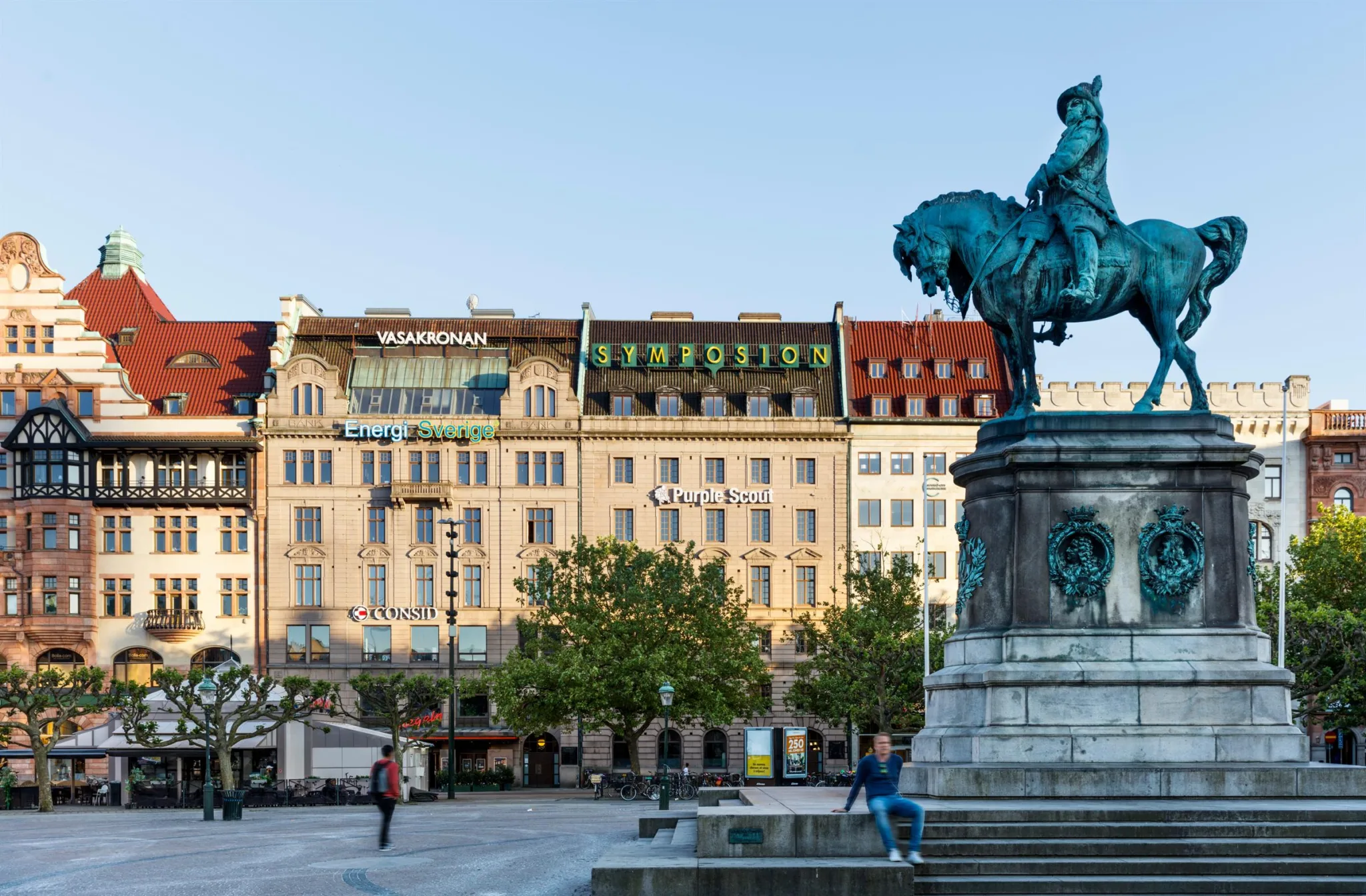 Staty av en ryttare på häst framför en bred byggnadsfasad med skyltar och träd i förgrunden.