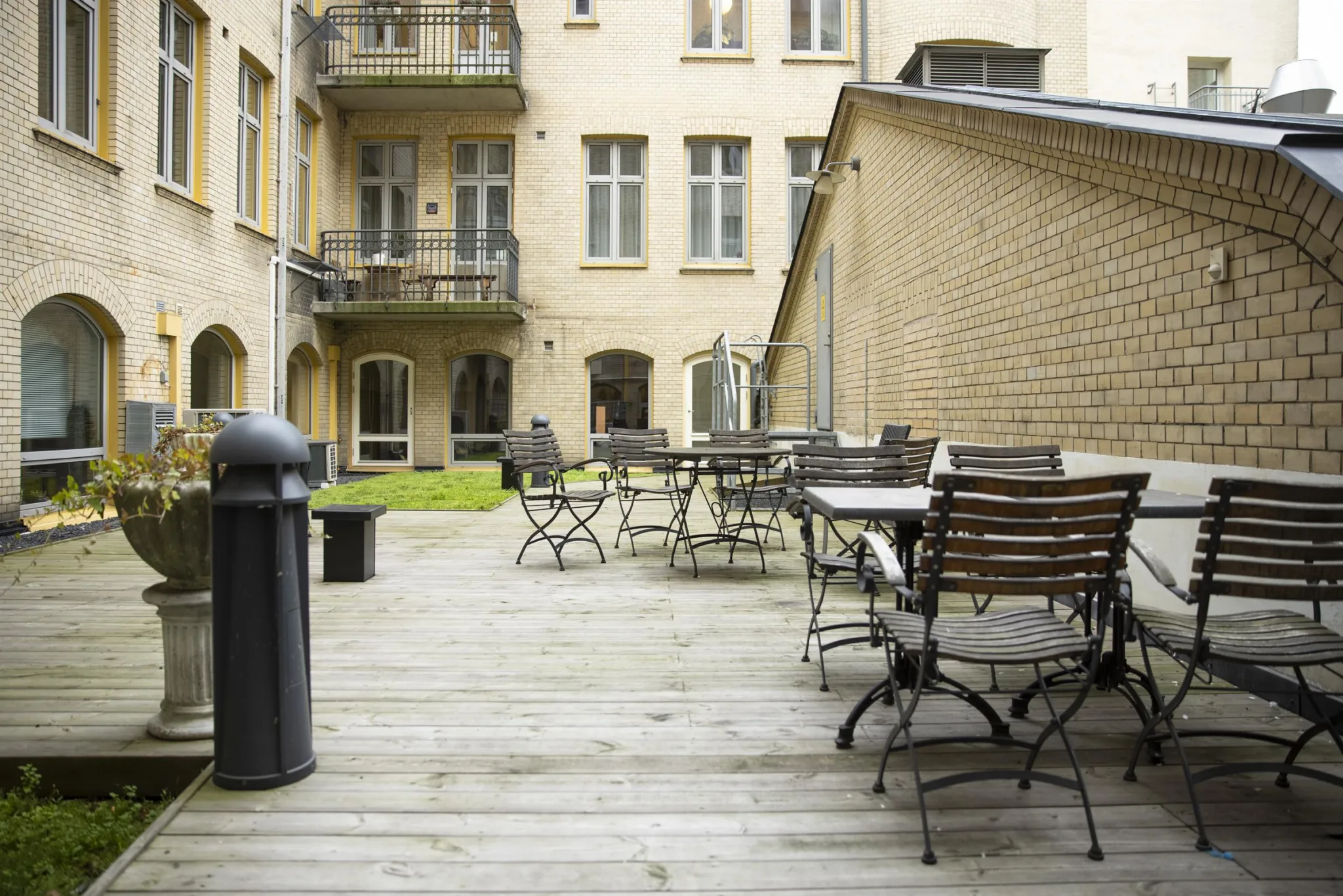 En innergård med trätrall, flera tomma cafébord och stolar, omgiven av beige byggnader.