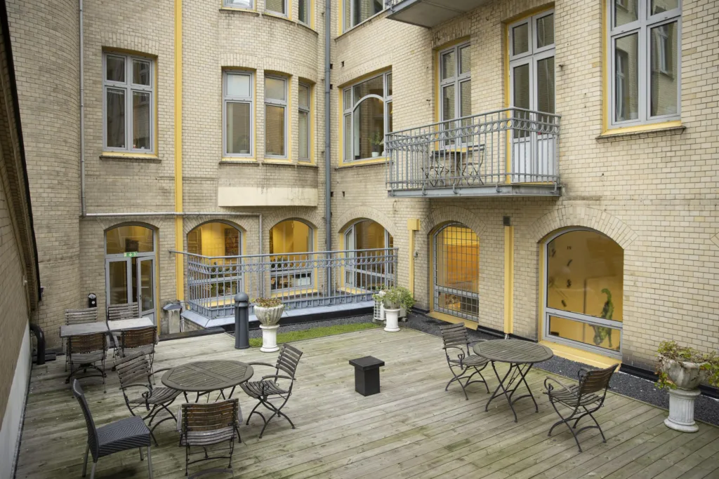 Innergård med bord, stolar och planteringar, omgiven av ljusa byggnader med fönster och balkonger.