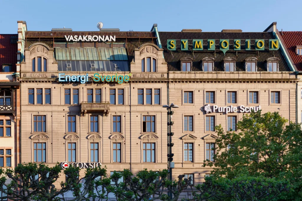En klassisk byggnad med skyltar som Vasakronan och Symposion på fasaden, omgiven av gröna träd.