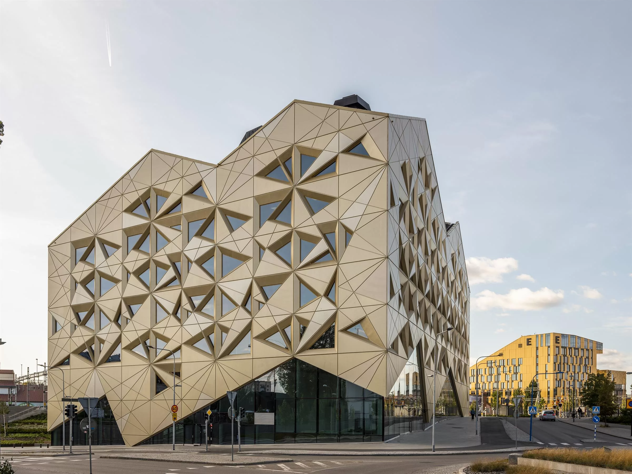 Modern byggnad med geometriska trianglar och glasfasad, belägen vid en väg med andra byggnader i bakgrunden.