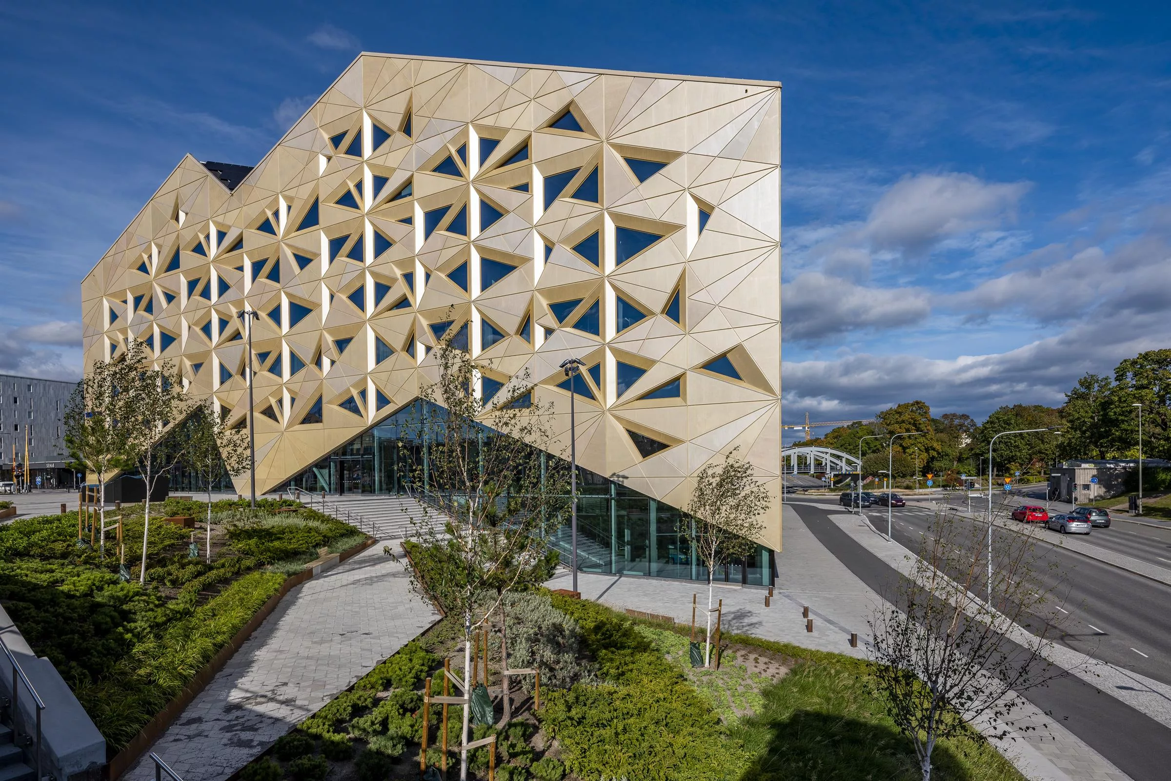 Modern byggnad med triangulär fasad och glas, omgiven av grönska och gångvägar, under en klarblå himmel.