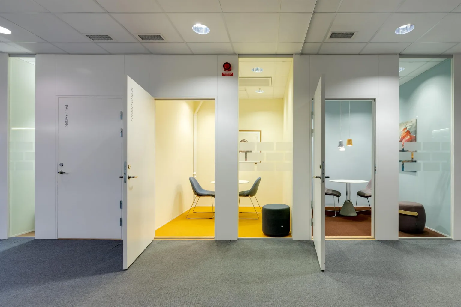 1. Två öppna dörrar som leder till små mötesrum med minimalistisk inredning; det ena rummet har en gul matta och det andra en grå matta.