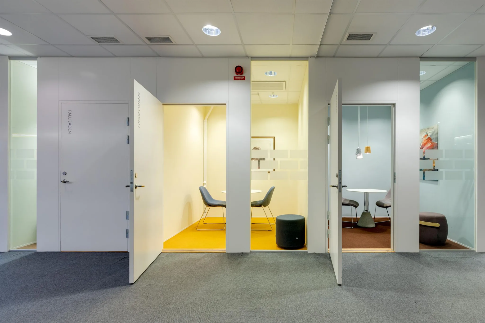 1. Två öppna dörrar som leder till små mötesrum med minimalistisk inredning; det ena rummet har en gul matta och det andra en grå matta.