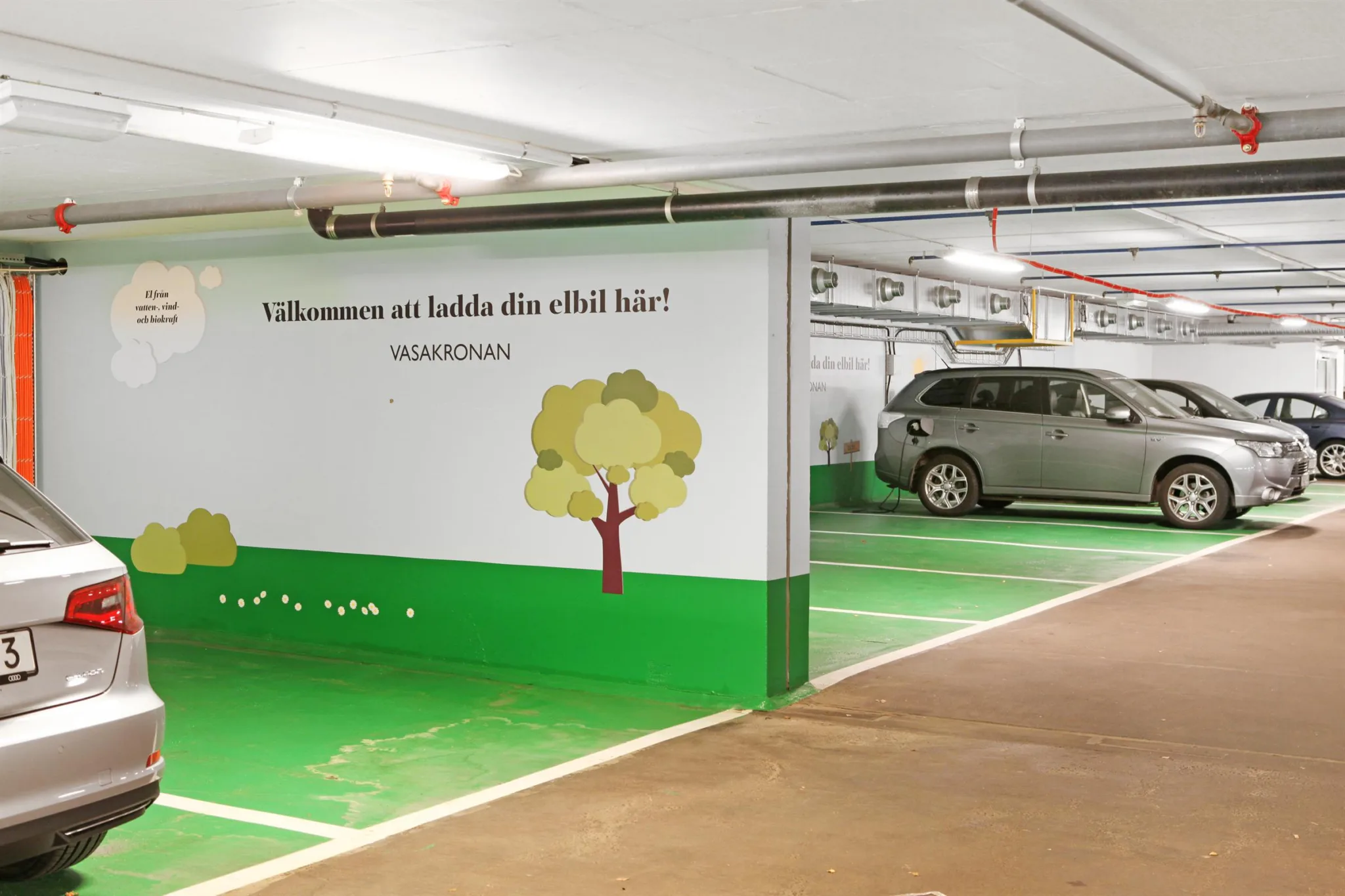 En parkeringsplats med en elbil och text på väggen som lyder: "Välkommen att ladda din elbil här! Hammarby." Väggmålningar av träd och gräsmattor.