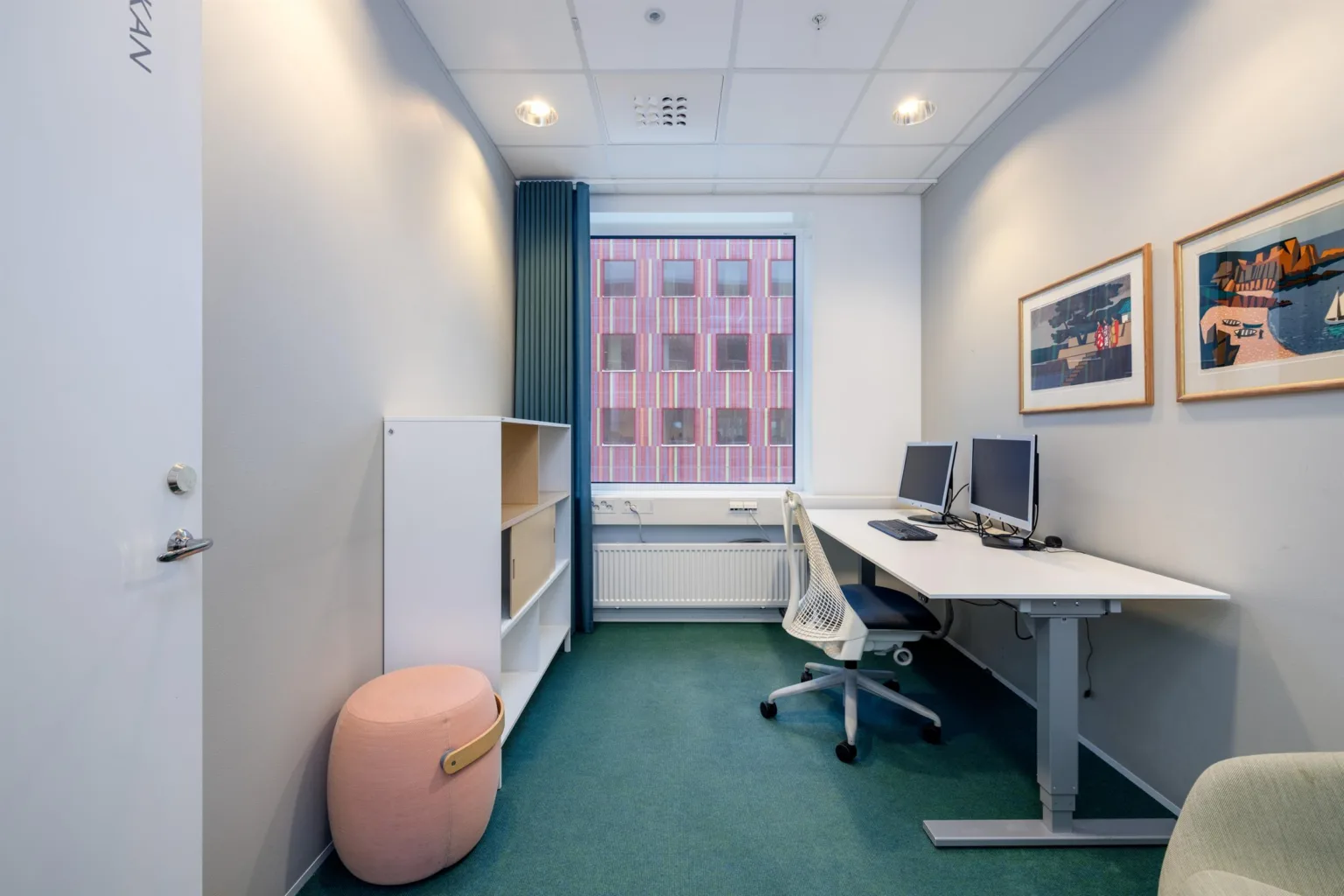 Ett mindre kontorsrum med två skrivbord, datorskärmar, hyllor och en rosa sittpuff. Ett fönster visar utsikt mot en röd byggnad.