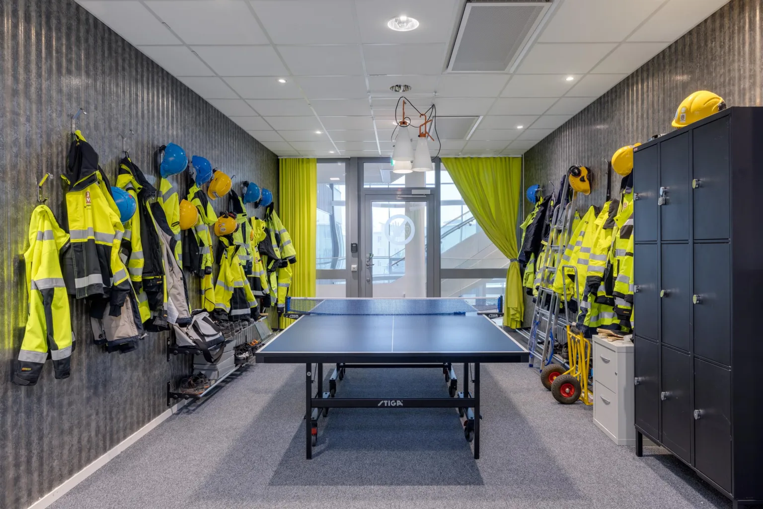 Ett rum med pingisbord i mitten, omgiven av skyddskläder och hjälmar på båda väggarna, med gröna gardiner vid ett fönster.