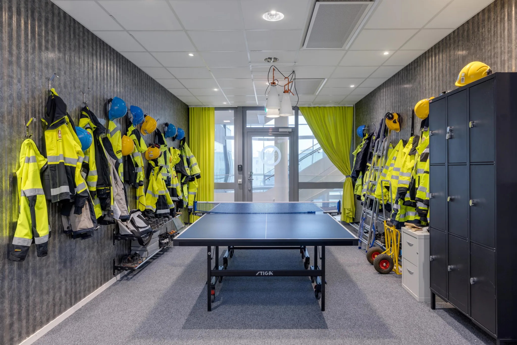 Ett rum med pingisbord i mitten, omgiven av skyddskläder och hjälmar på båda väggarna, med gröna gardiner vid ett fönster.