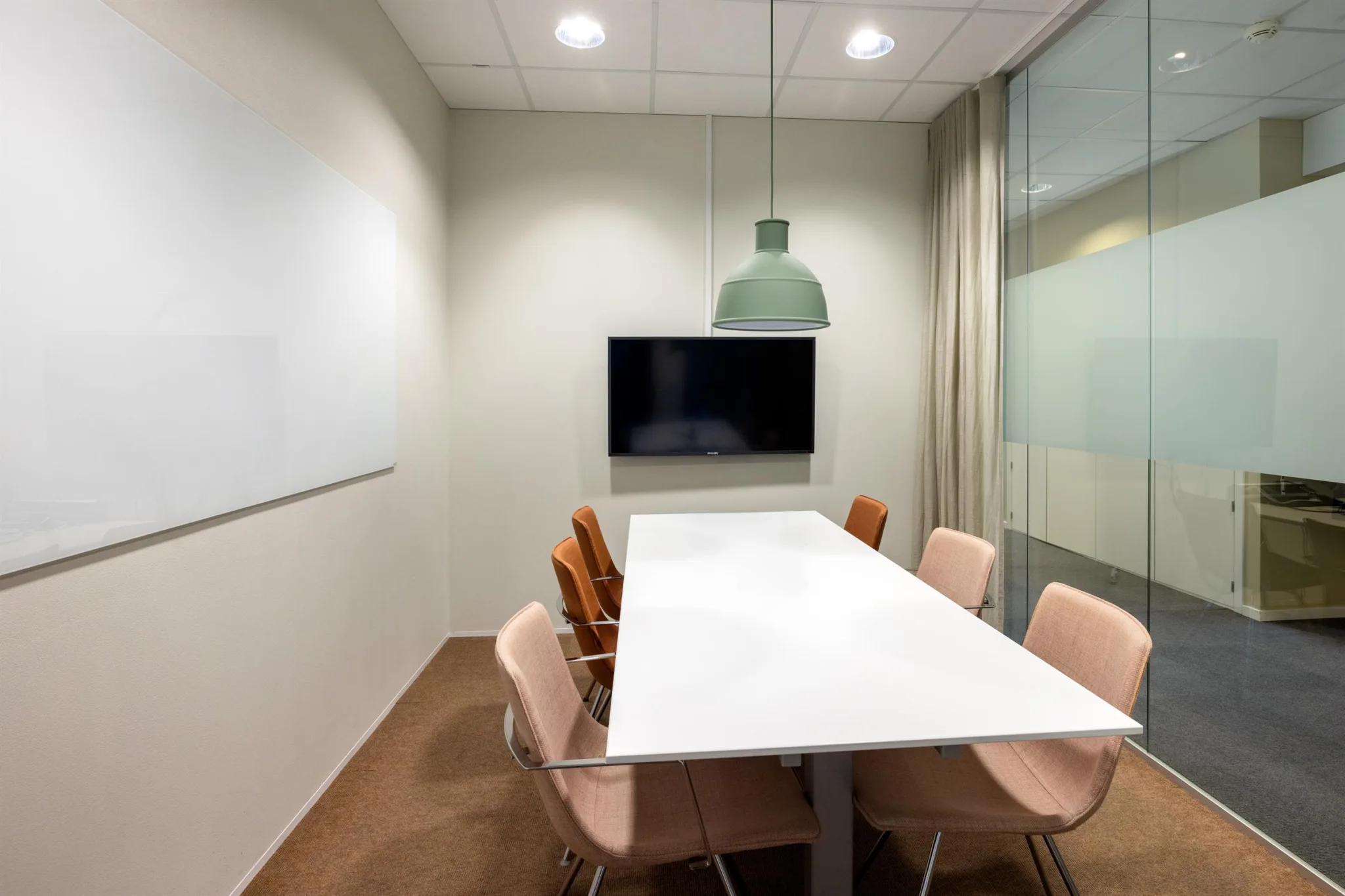 Ett konferensrum med ett vitt bord, rosa stolar, en väggmonterad skärm och en grön taklampa.
