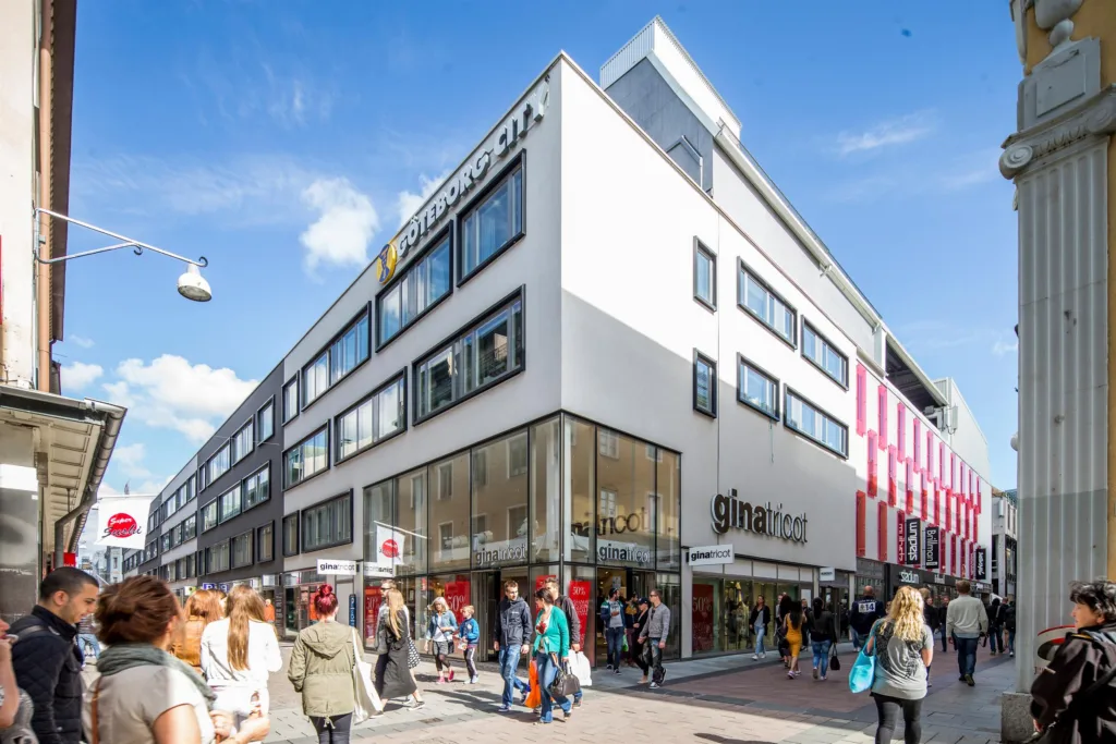 En vit byggnad med stora glasfönster och skylten "gina tricot", omgiven av människor som promenerar under en blå himmel.