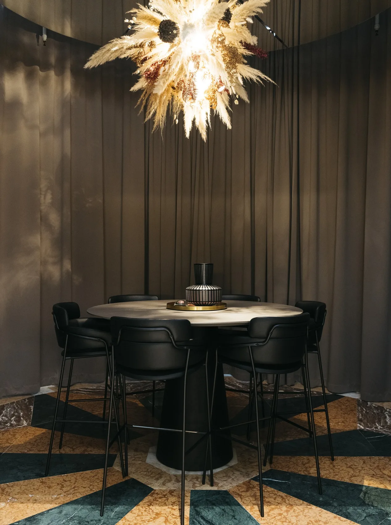 Matbord med svarta stolar och en modern lampa med fjädrar i taket, omgiven av mörka gardiner.