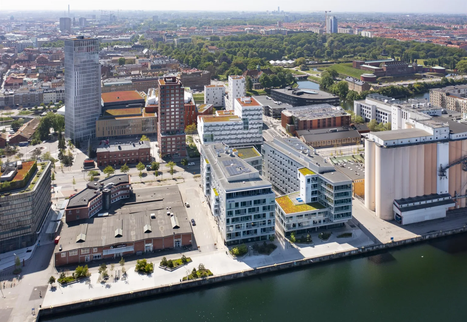 Flygfoto över ett modernt stadsområde med höga byggnader och grönområden vid en kanal.