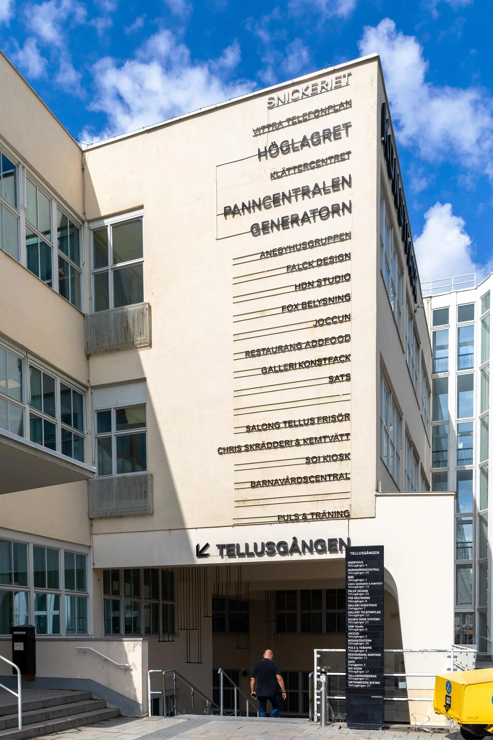 4. Vit byggnad med text på fasaden som listar olika verksamheter, blå himmel och glasdetaljer i bakgrunden.