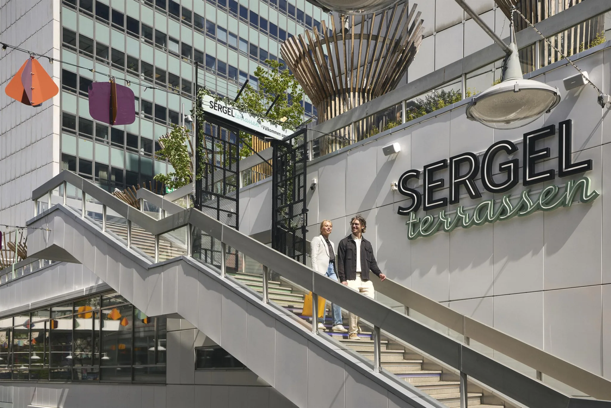5. Två personer går nerför en trappa utanför Sergels torg, med byggnadsskyltar och ett konstverk i bakgrunden.