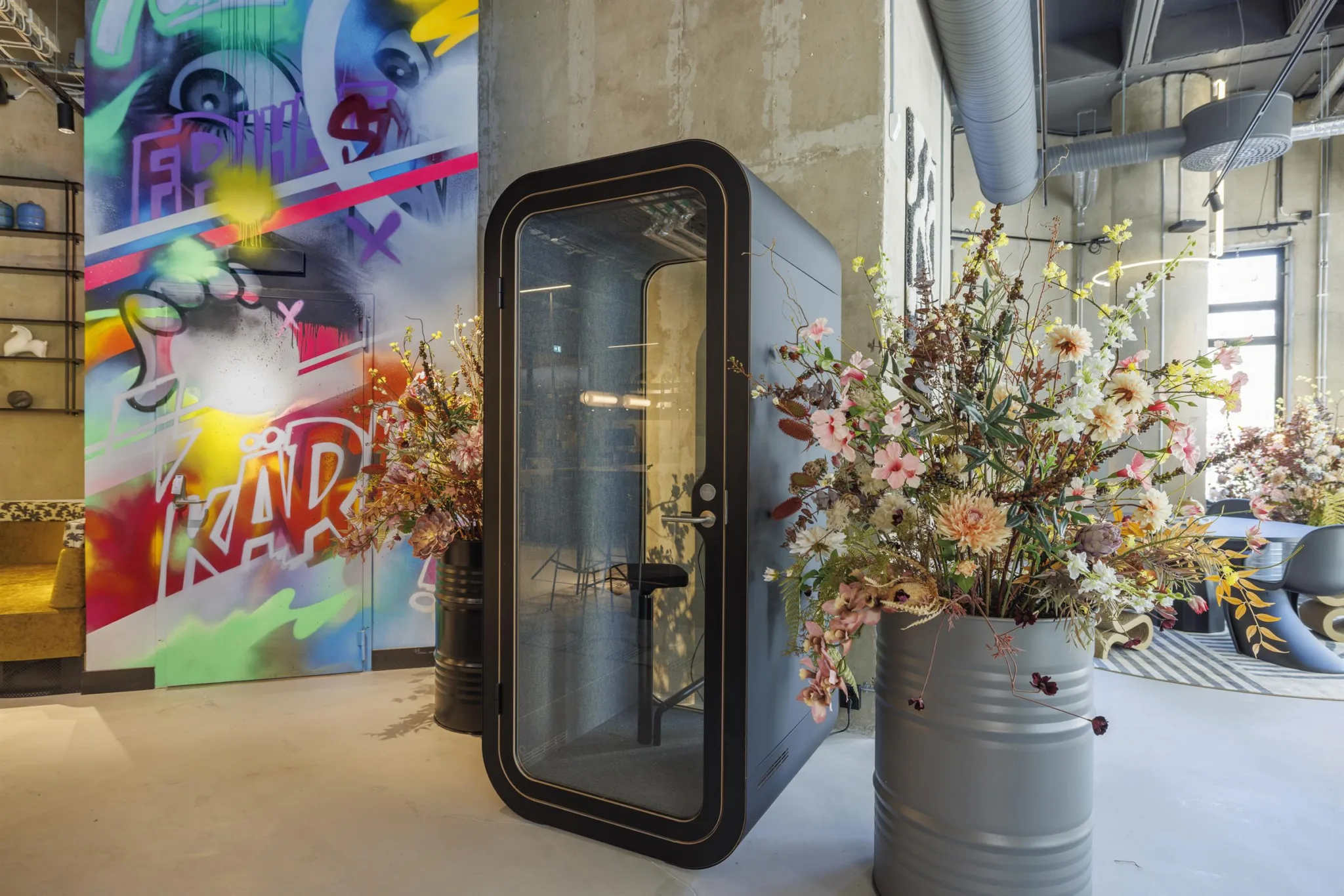 En ljudisolerad telefonkabin bredvid blomsterarrangemang i metallkrukor, med en färgglad väggmålning i bakgrunden.