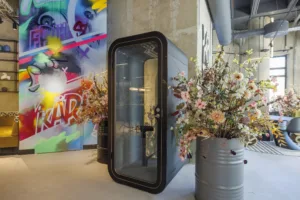 En ljudisolerad telefonkabin bredvid blomsterarrangemang i metallkrukor, med en färgglad väggmålning i bakgrunden.