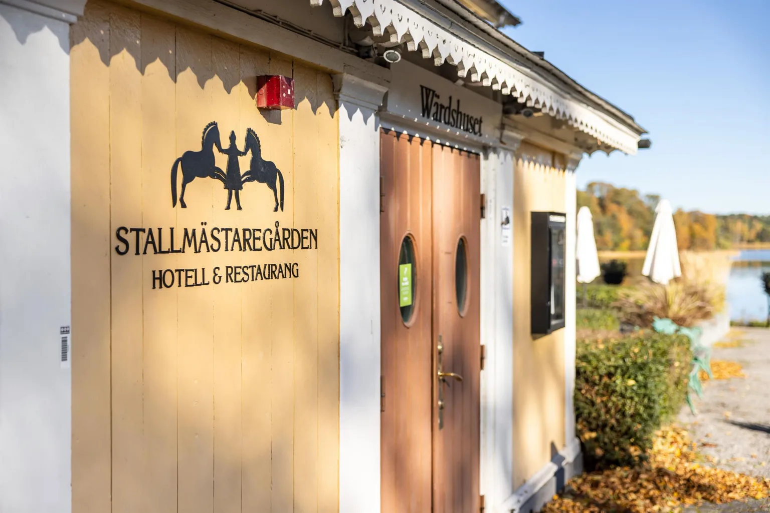 2. Gul byggnad med texten "TALLMÄSTAREGÅRDEN HOTELL & RESTAURANG" på väggen, en träport och omgivande grönska.
