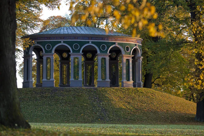 1. En paviljong med valvbågar står på en grön kulle omgiven av träd med höstfärger.
