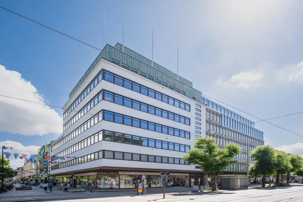 1. En modern byggnad med vita fasader och stora fönster, omgiven av ett träd och en trottoar under en klarblå himmel.