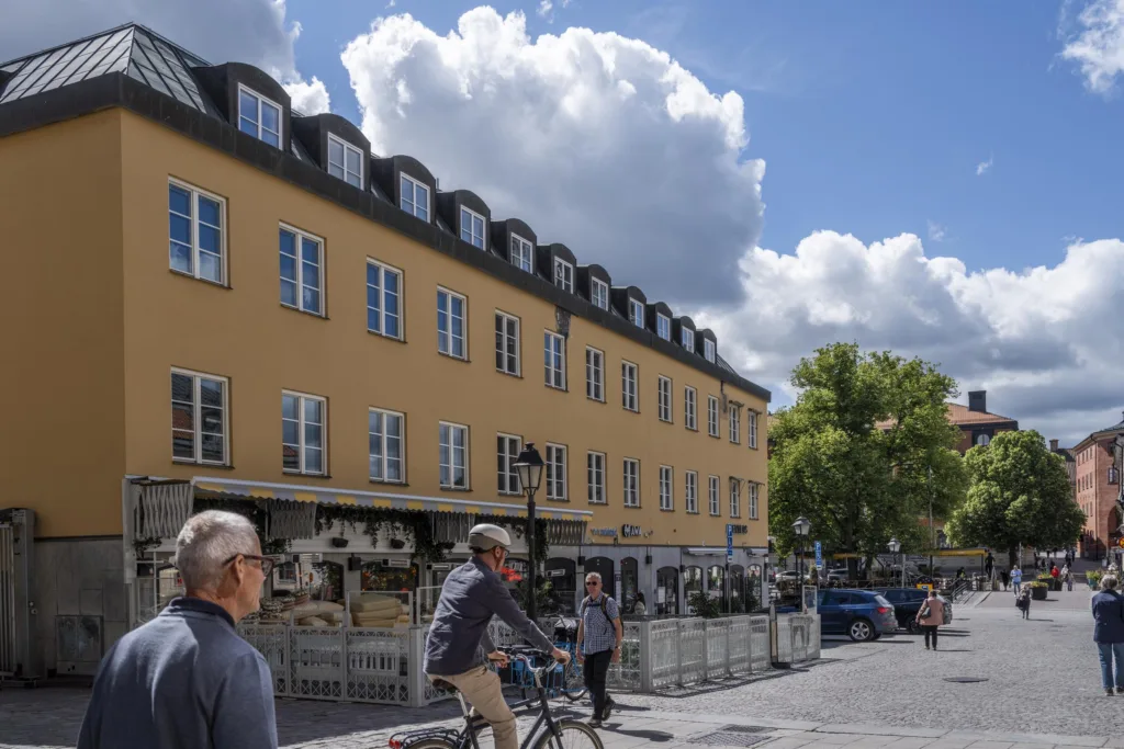 En gul byggnad med uteservering, människor går och cyklar på en solig gata med moln på himlen.