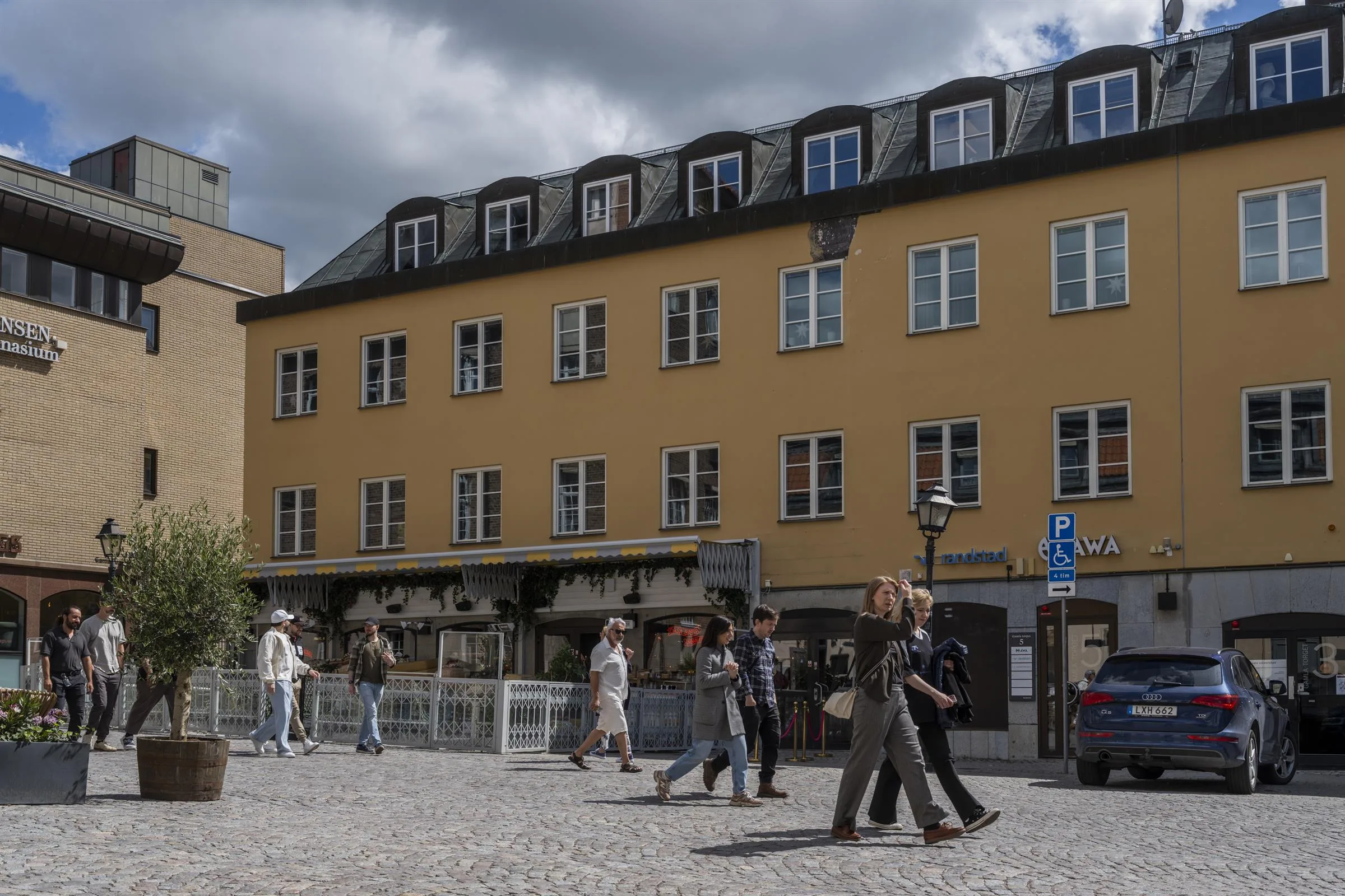 En gul byggnad med flera fönster, människor promenerar på en stenlagd gata under molnig himmel.