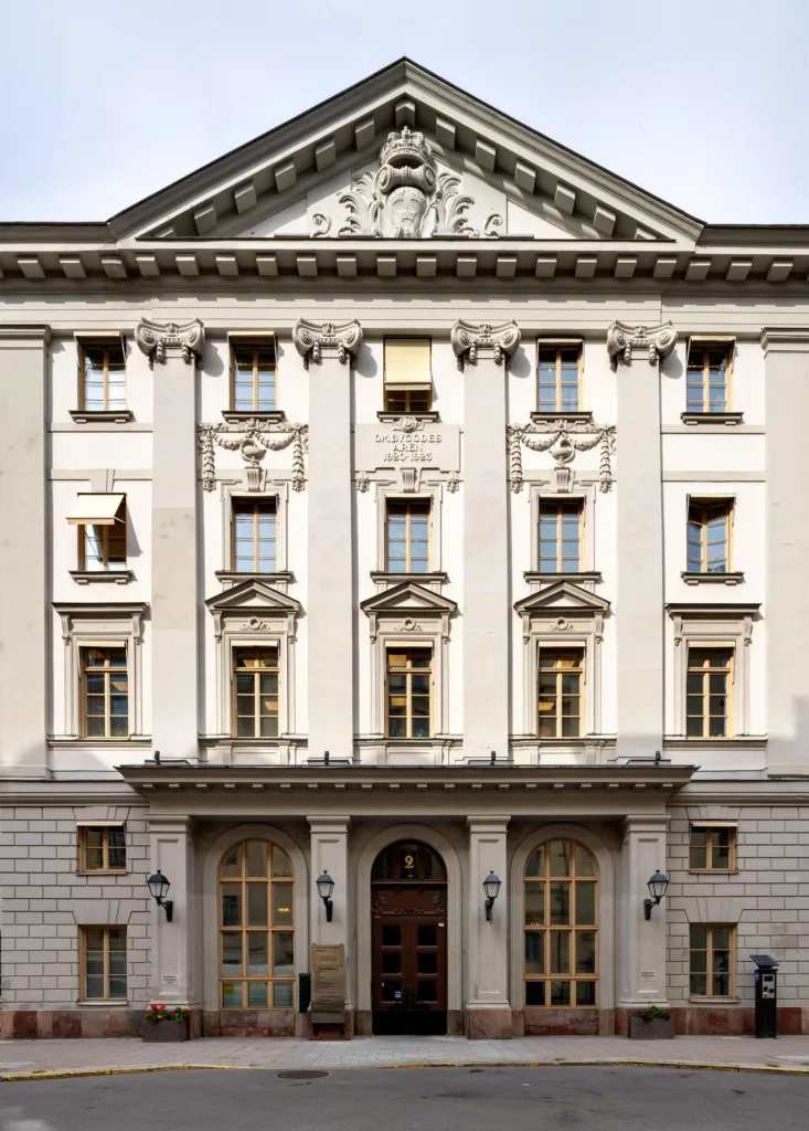 En vit byggnad med klassisk arkitektur, flera fönster och en välvd entré med dekorativa detaljer.