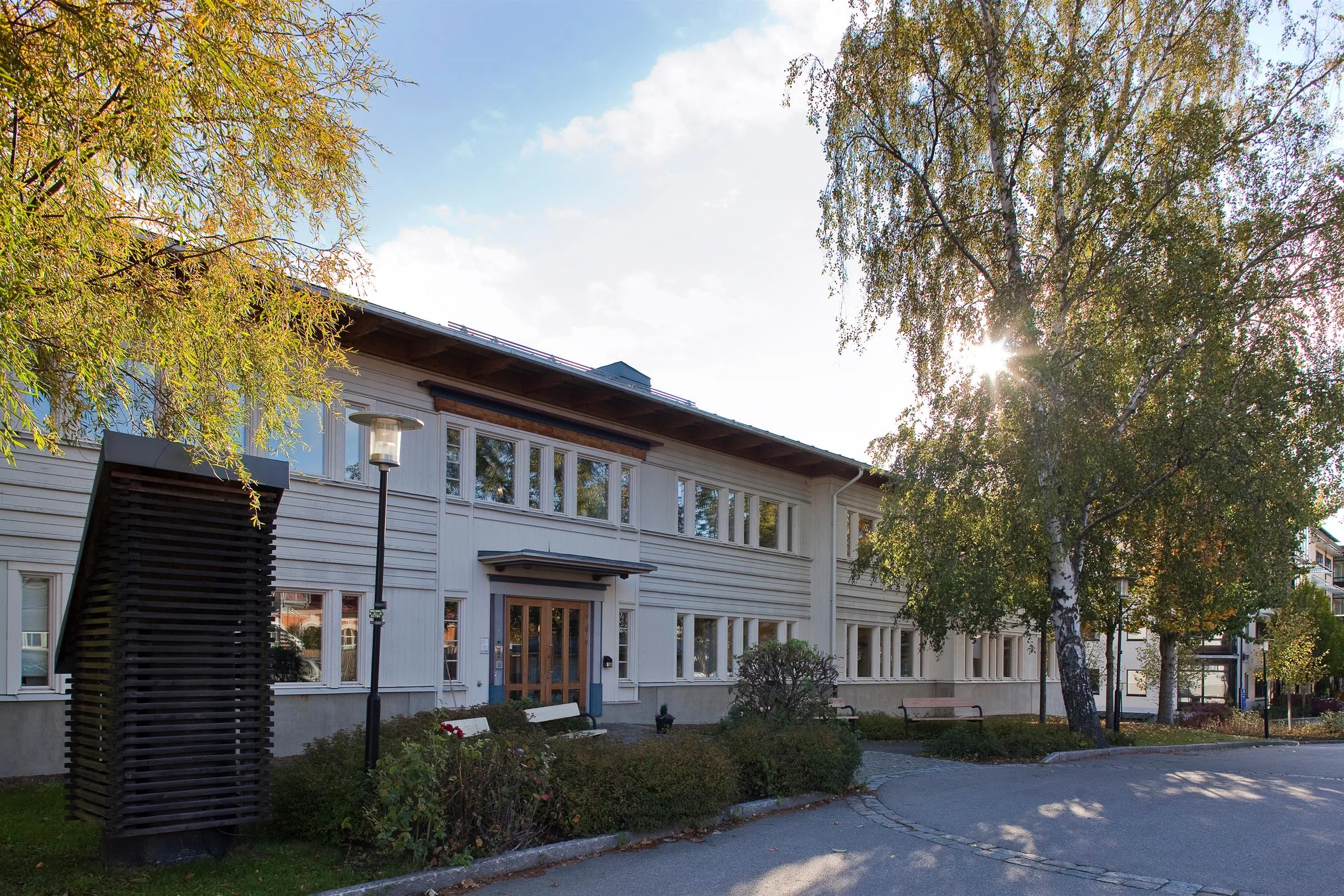 En vit byggnad med trädetaljer, omgiven av träd och ett upplyst område i dagsljus.