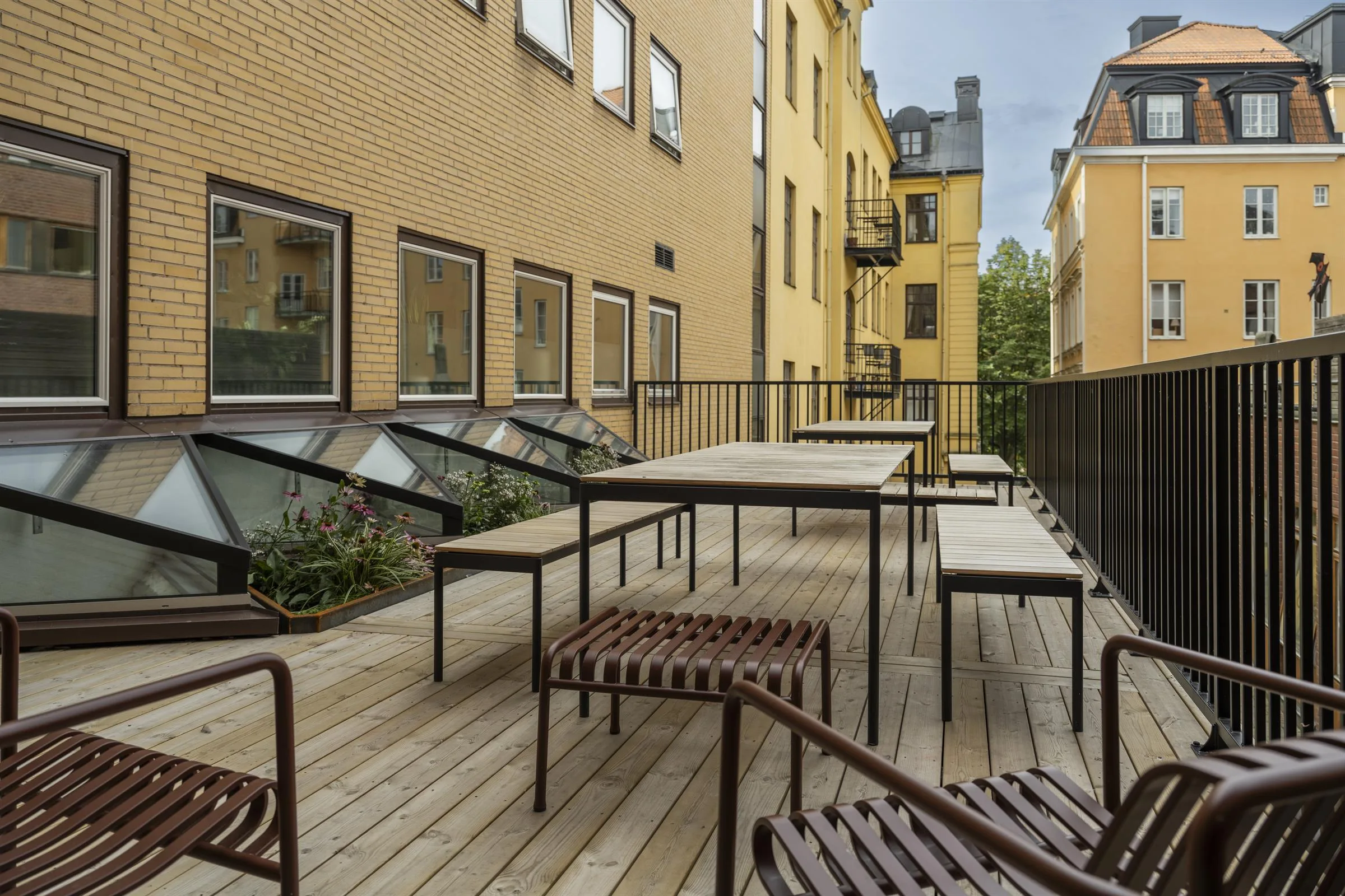 2. En uteplats med träbord och metallstolar omgiven av gula byggnader och grönska.