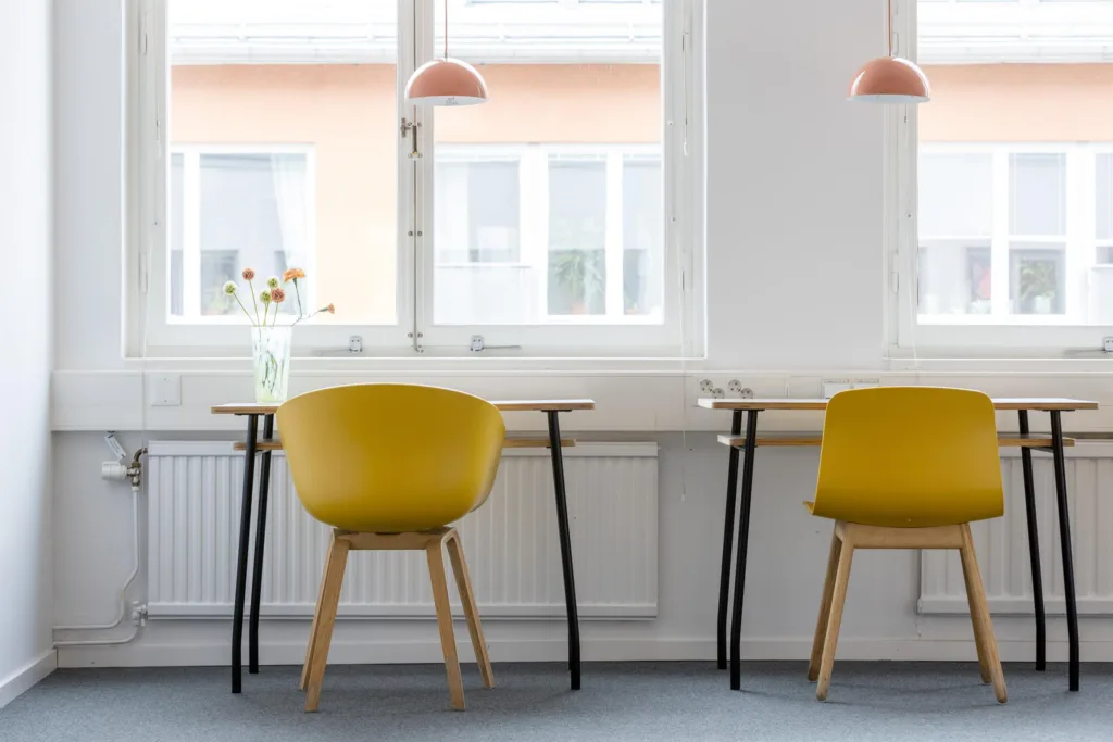 Två skrivbord med gula stolar, rosa taklampor och fönster med utsikt mot byggnader.