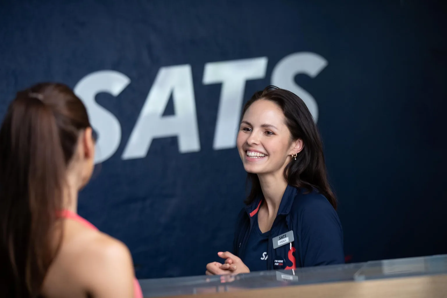 5. En leende kvinna i en mörkblå uniform står vid en disk med texten "SATS" på väggen bakom.