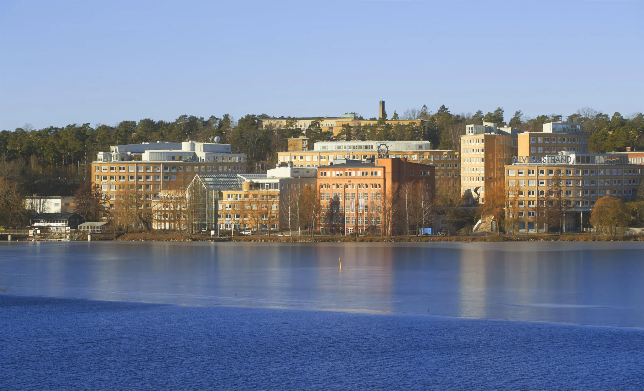 En vy över flera byggnader på en kulle, omgivna av träd och med en sjö i förgrunden.