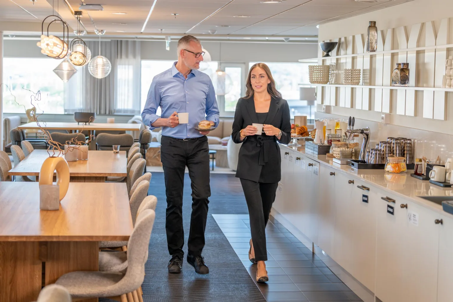 En man och en kvinna går genom ett kontorskök med kaffekoppar i händerna.
