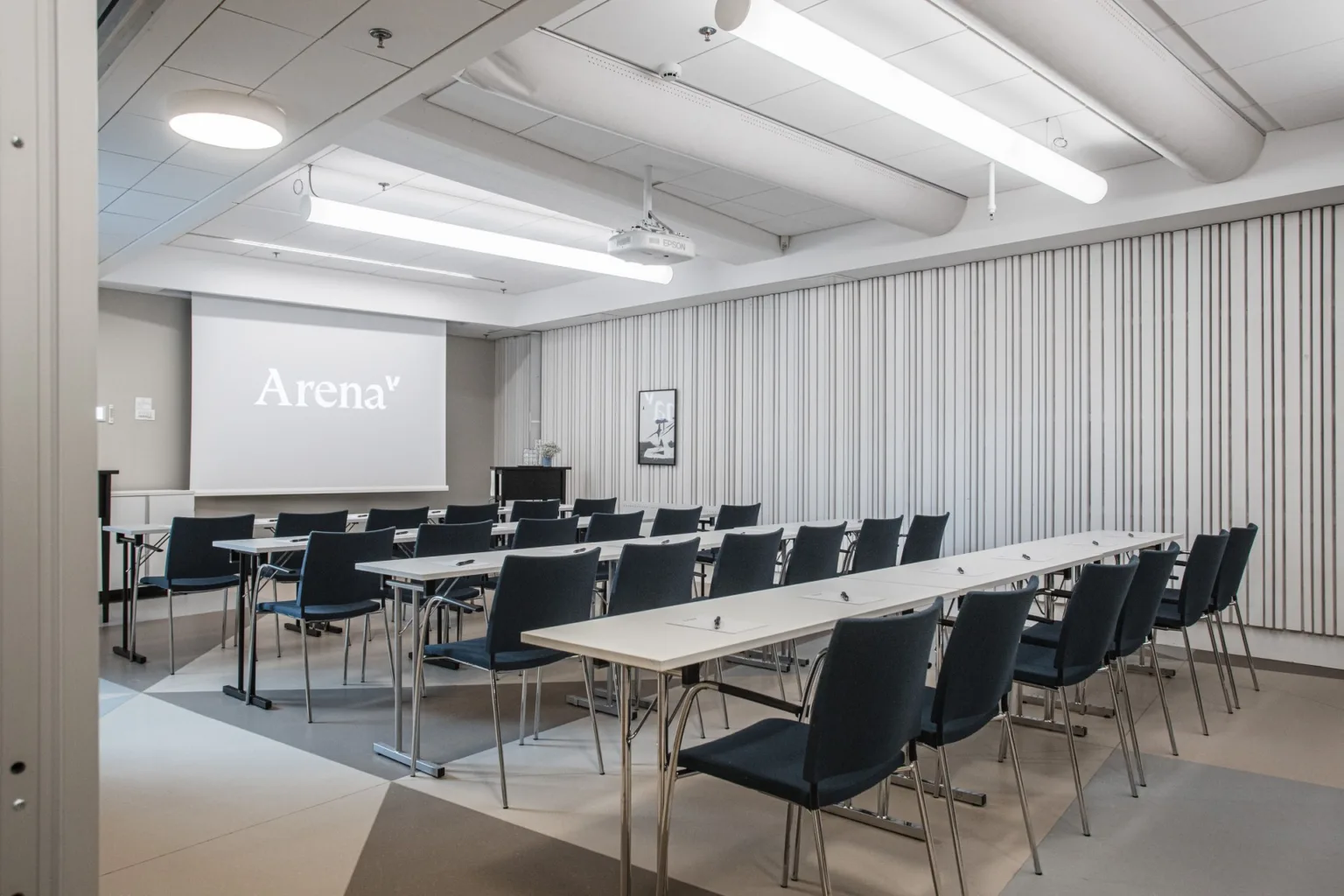 En konferenssal med vita bord och svarta stolar, en projektorduk med texten "Arena" samt dämpad belysning.