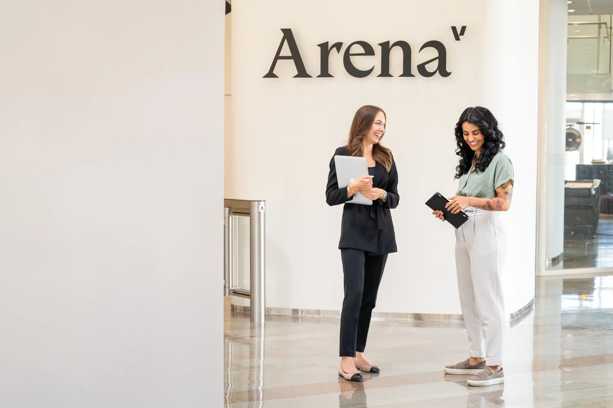 Två kvinnor står framför en vägg med texten "Arena"; en håller dokument och den andra håller en telefon.