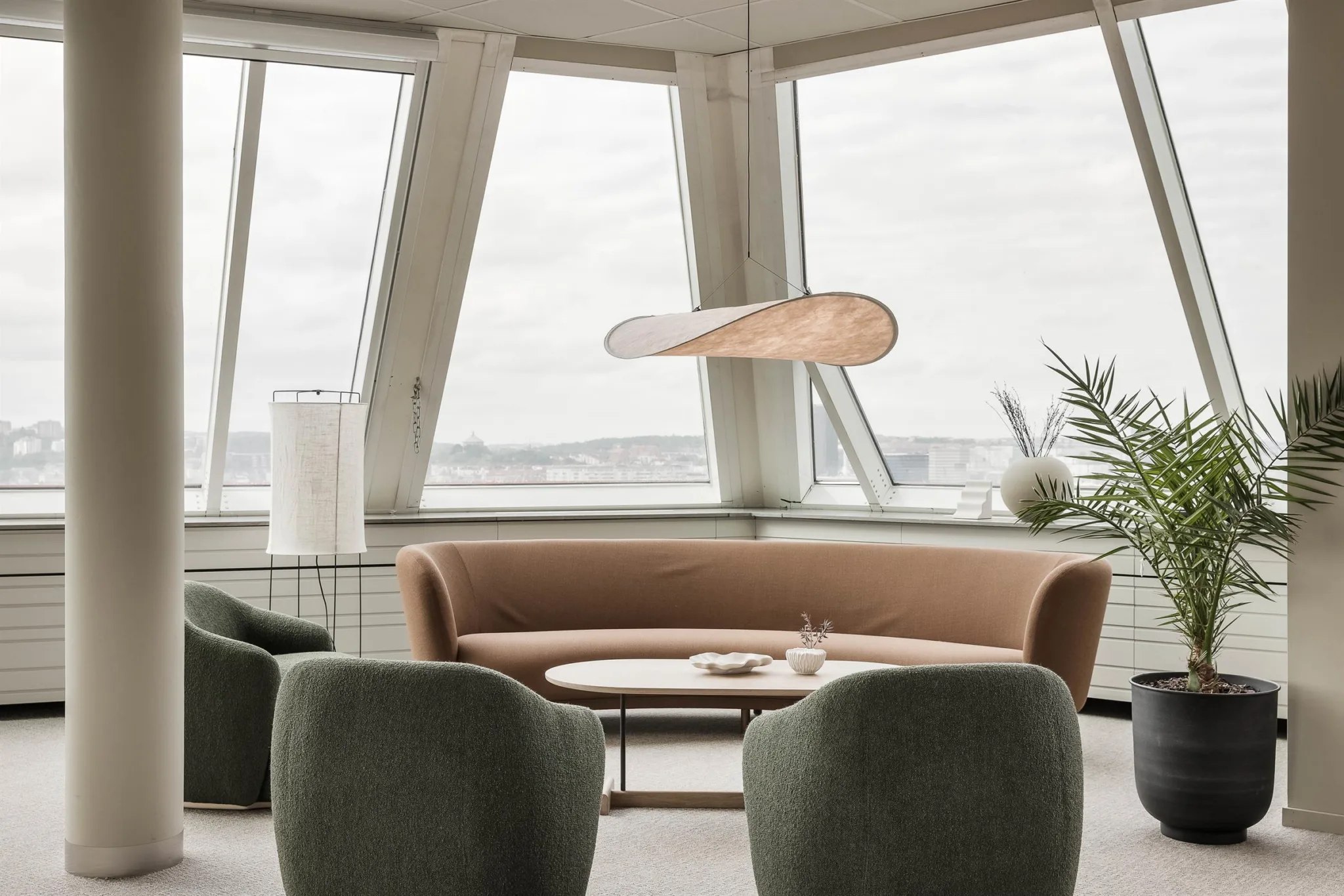 En stilren lounge med en beige soffa, gröna fåtöljer och ett modernt hängande ljus framför stora fönster.