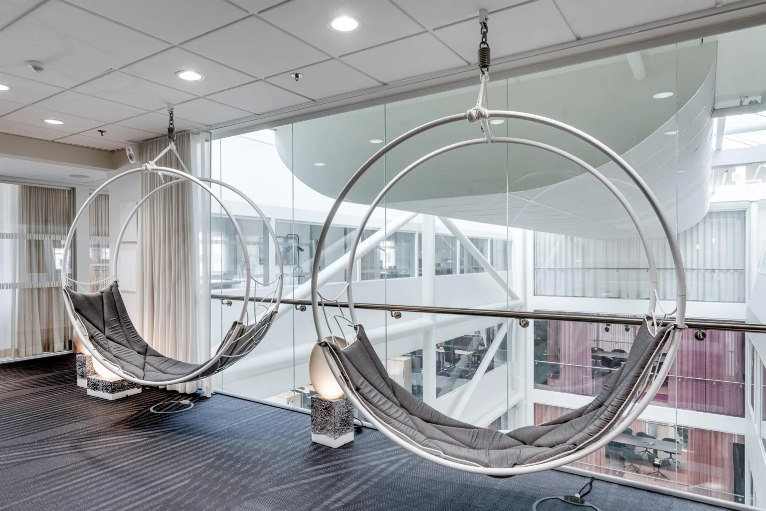 Två hängande stolar med metallramar och tygsitsar i en ljus och modern inomhusmiljö med glasväggar.