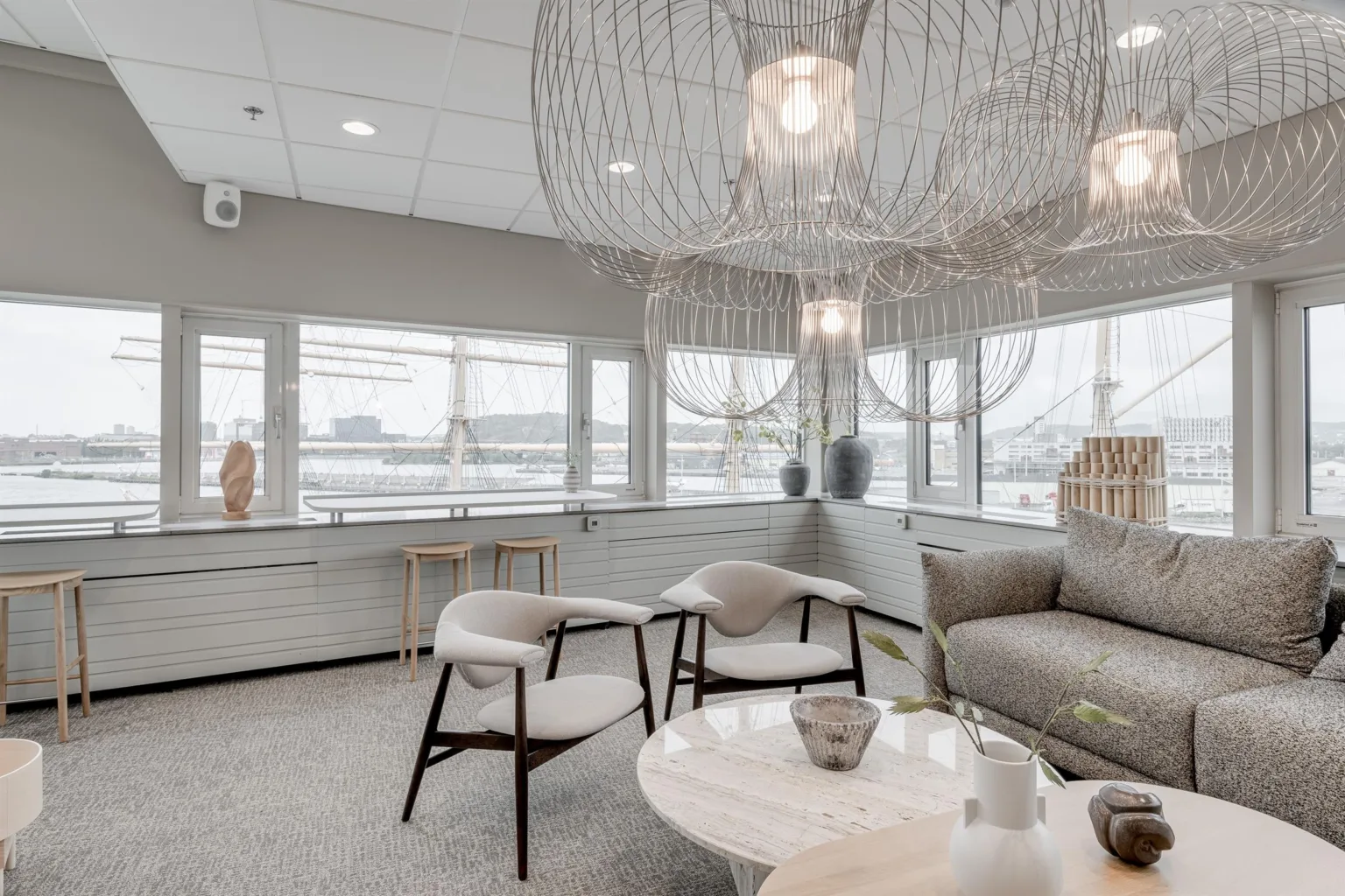 Ett ljust rum med bekväma stolar, en soffa, ett runt bord och dekorativa taklampor, samt fönster som släpper in mycket naturligt ljus.