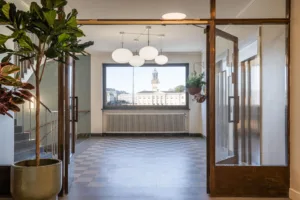 En korridor med glasdörrar, stora fönster och hängande taklampor, med utsikt över ett kyrktorn i fjärran.