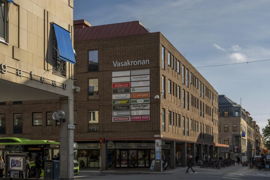 Brun tegelbyggnad med texten "Vasakronan" och fönster täckta med olika färgade skyltar.