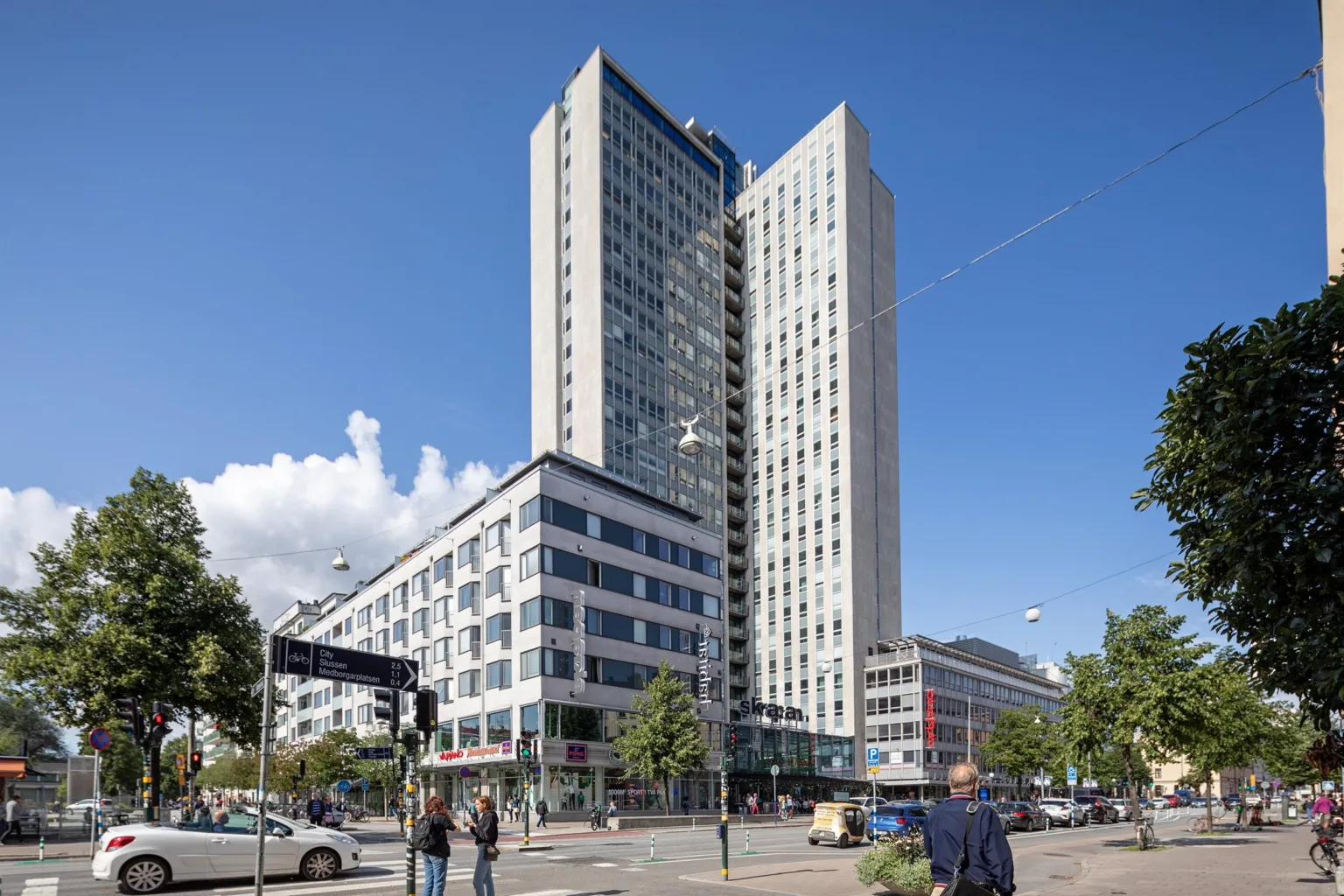 En hög modern byggnad med glasfasad och kringliggande stadsmiljö under en klarblå himmel.