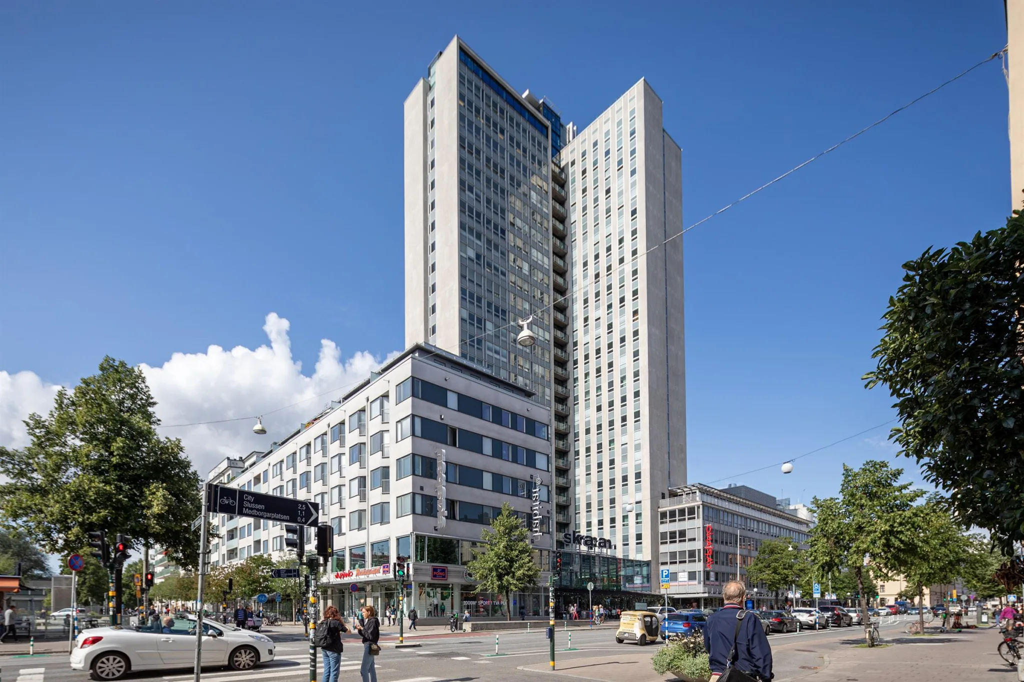 En hög modern byggnad med glasfasad och kringliggande stadsmiljö under en klarblå himmel.