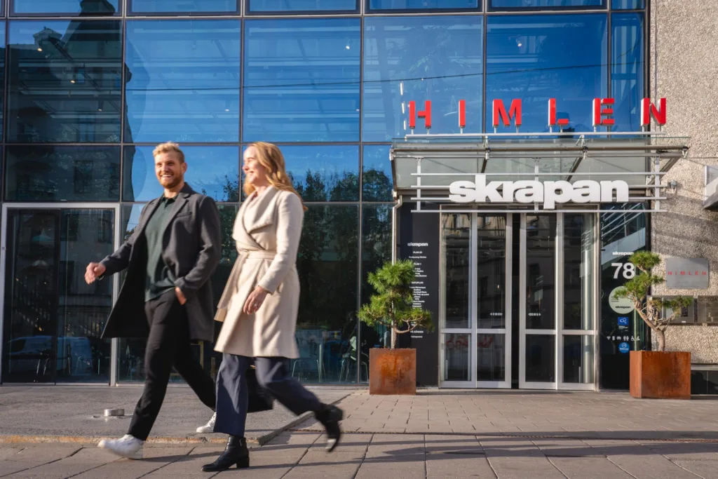 Två personer promenerar framför en byggnad med glasfasad, där texten "Himlen Skrapan" syns ovanför ingången.