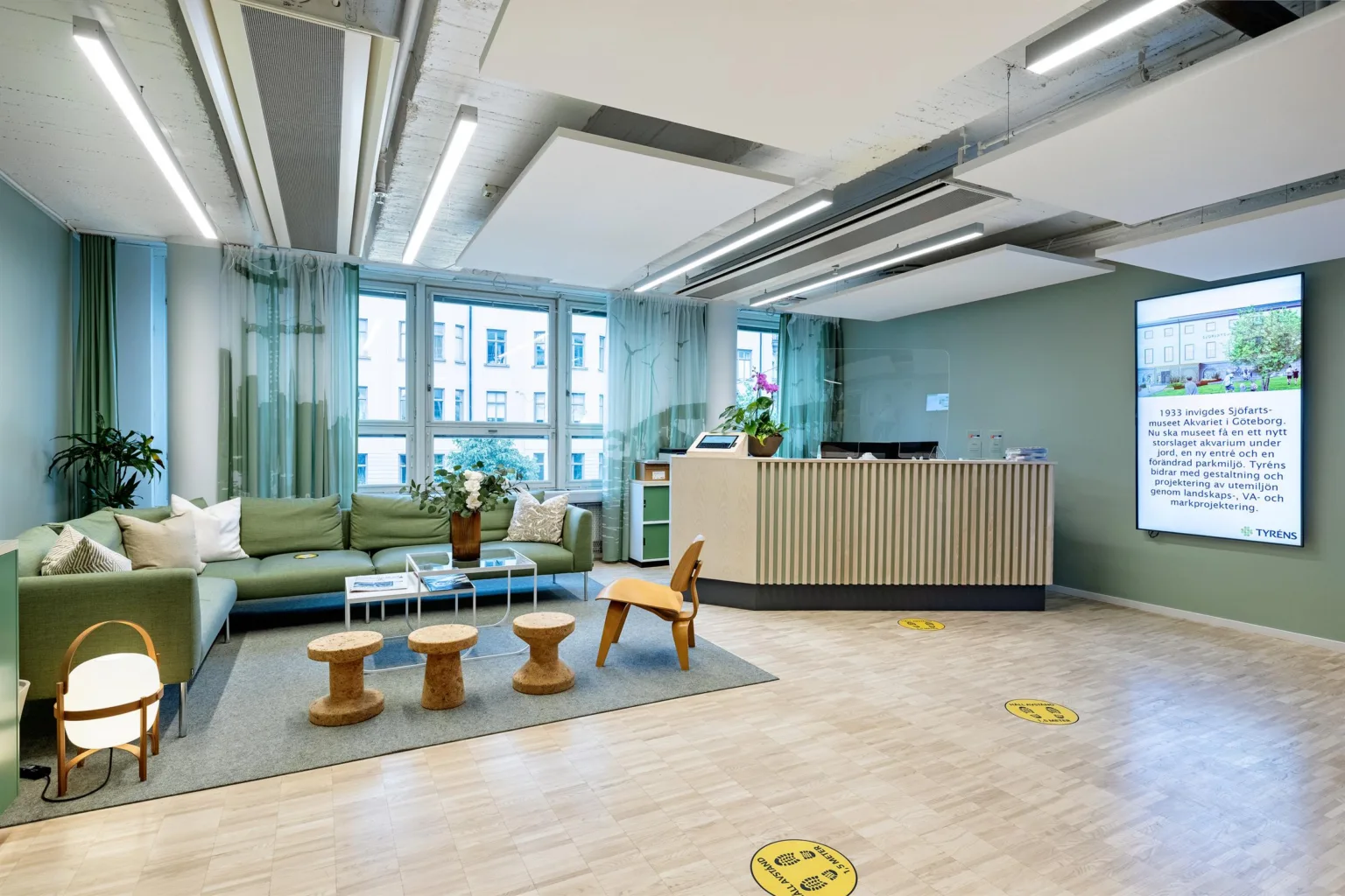 En receptionsyta med en soffa, små bord och en träleksakshäst, omgiven av gröna väggar och stora fönster.