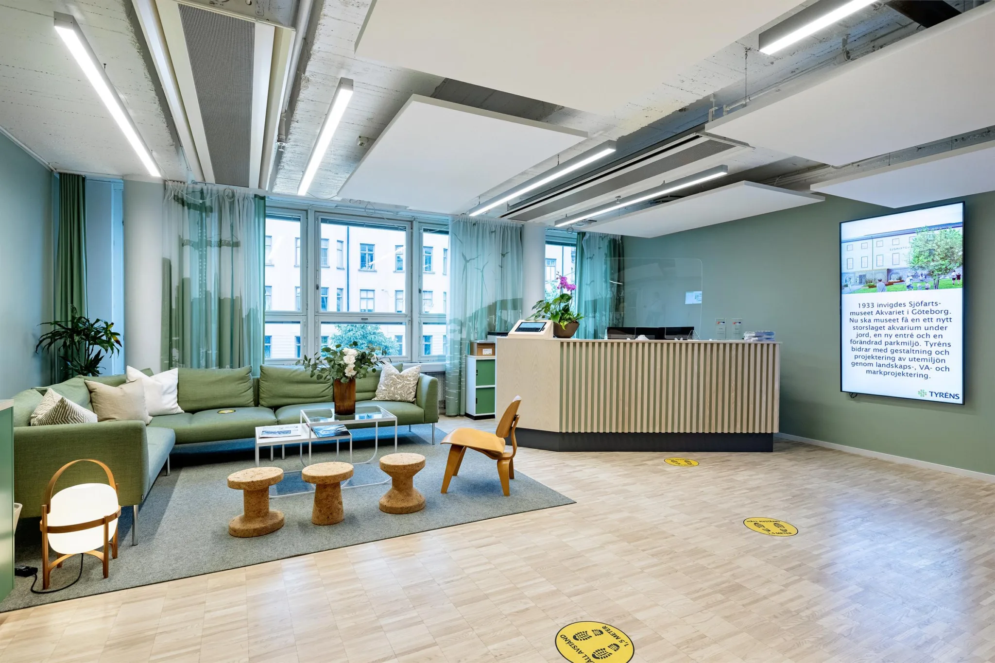 En receptionsyta med en soffa, små bord och en träleksakshäst, omgiven av gröna väggar och stora fönster.