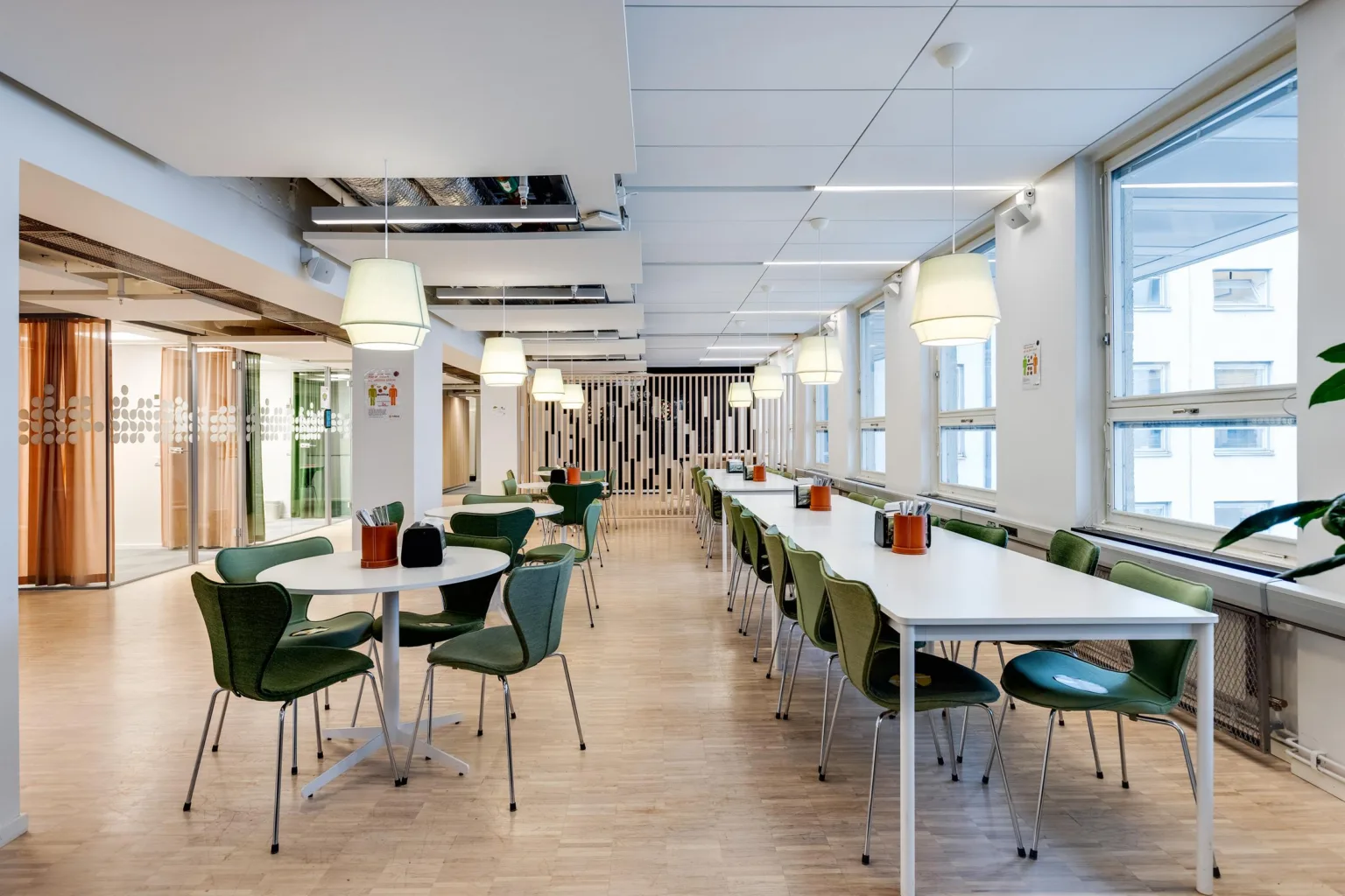 En matsal med vita bord, gröna stolar och stora fönster, inredd med moderna lampor och växter.