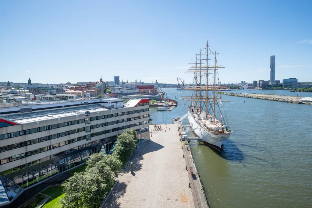 En utsikt över en hamn med ett stort segelfartyg, byggnader och en promenad vid vattnet under en klarblå himmel.
