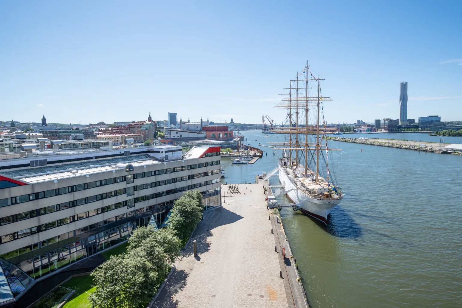 En utsikt över en hamn med ett stort segelfartyg, byggnader och en promenad vid vattnet under en klarblå himmel.